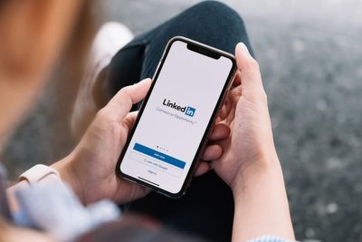 A person is looking down at their iPhone and reading the screen which shows the LinkedIn logo and login screen. You can Connect to Opportunity by being present on this platform with a LinkedIn business page.