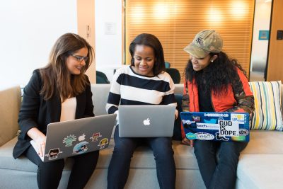Three women, two Black one white, sit side by side on a couch with their laptops on their laps. They are collaborating and working together discussing something while smiling.