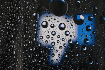 Raindrops cover a sheet of glass, sharply in focus. Each drop shows a Facebook thumbs up icon, seemingly reflected from a large, out of focus thumbs down icon in the background of the image.