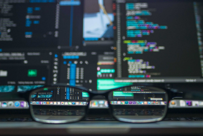 A pair of eyeglasses sit in the foreground in sharp clarity to the blurred background of a series of computer screens showing html script and coding script across the screens in blues and greens. 