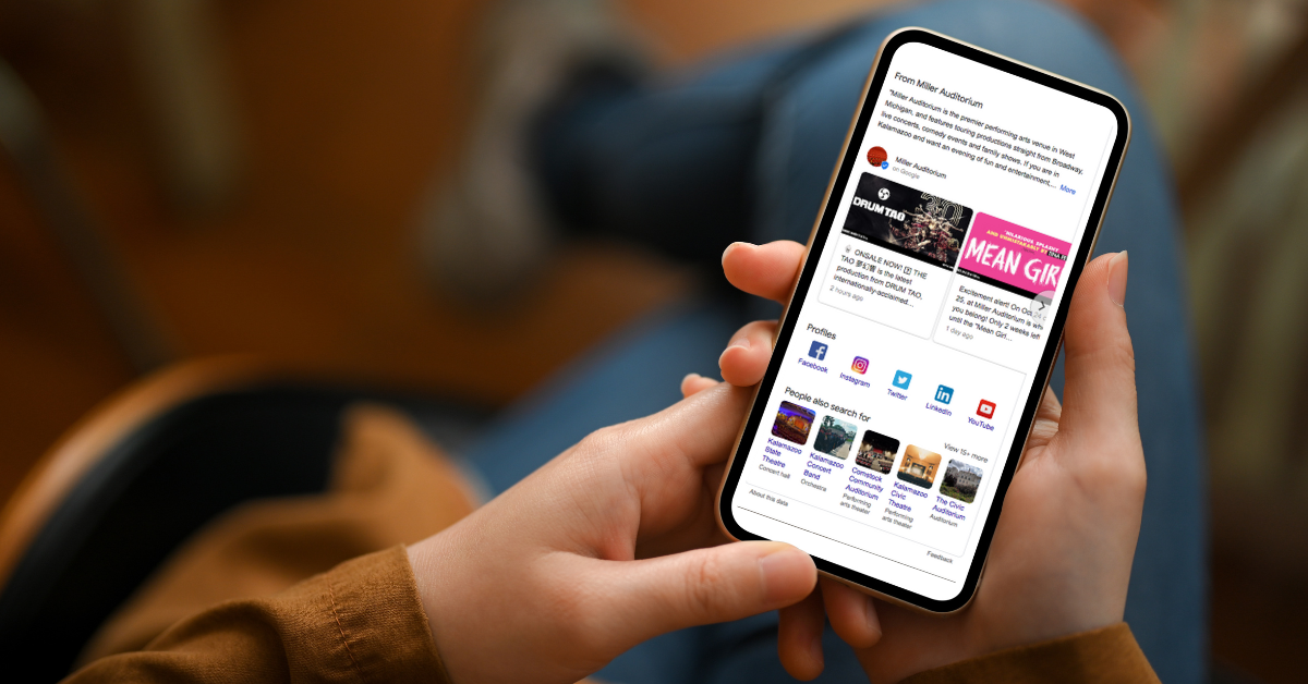 A person holding a smart phone with the phone in focus, the rest blurred. The phone face shows a Google Business Profile for Miller Auditorium, showing the icons for social media on the profile indicating this profile took advantage of the latest update.
