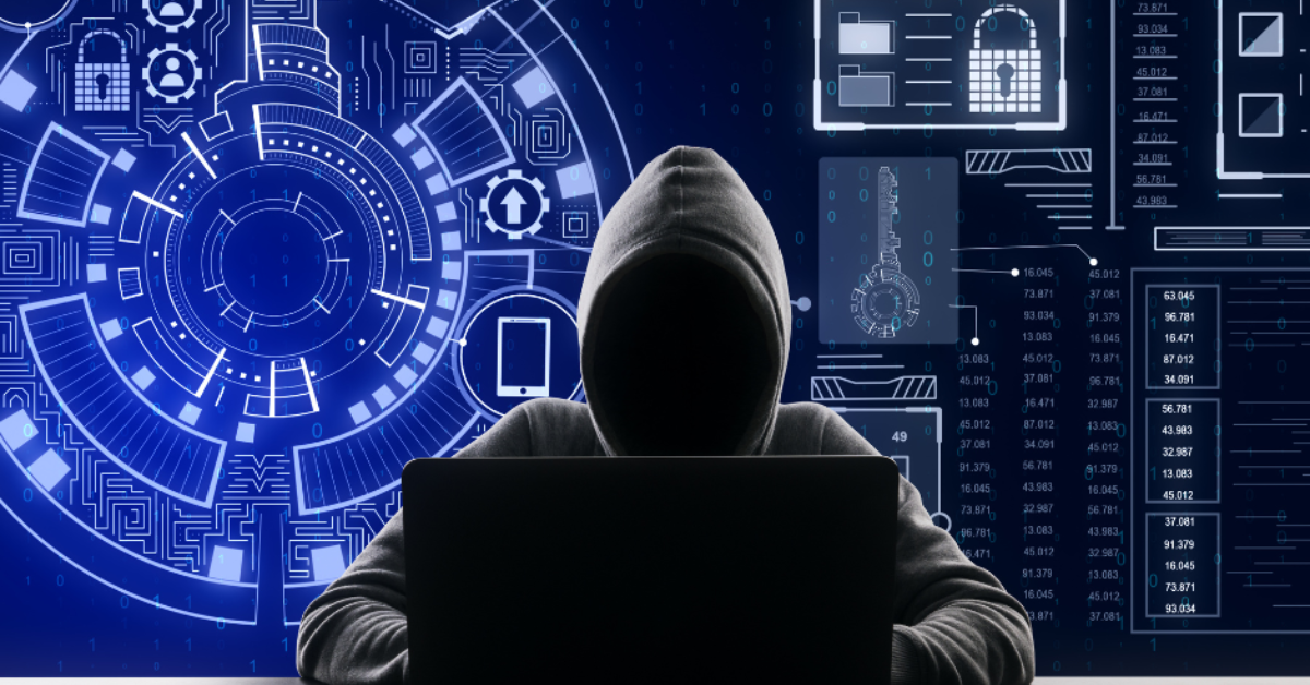 A person in a dark grey hooded sweatshirt sits with the hood up, face completely obscured, in front of an open laptop computer. Behind the person in the background is a digital screen in shades of blue with white writing and drawings and symbols about security such as locks.
