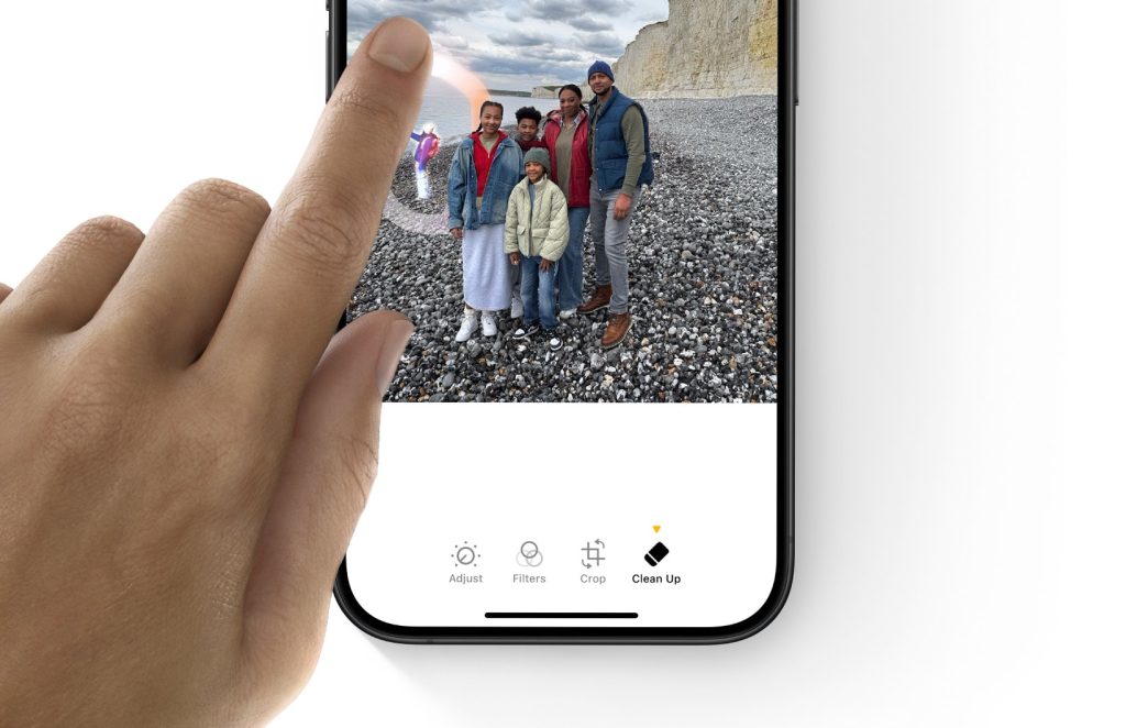 Shows a hand removing unwanted background items in an image on an iPhone 16 Pro by touching the image with their left pointer finger.