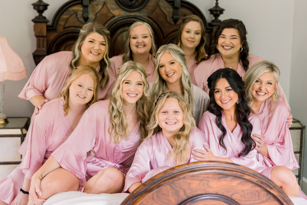 Wedding Party of bride and bridesmaids wearing silk pink robes and piled onto a bed inside Solomon Fowler Mansion featuring an ornate wooden headboard and foot board